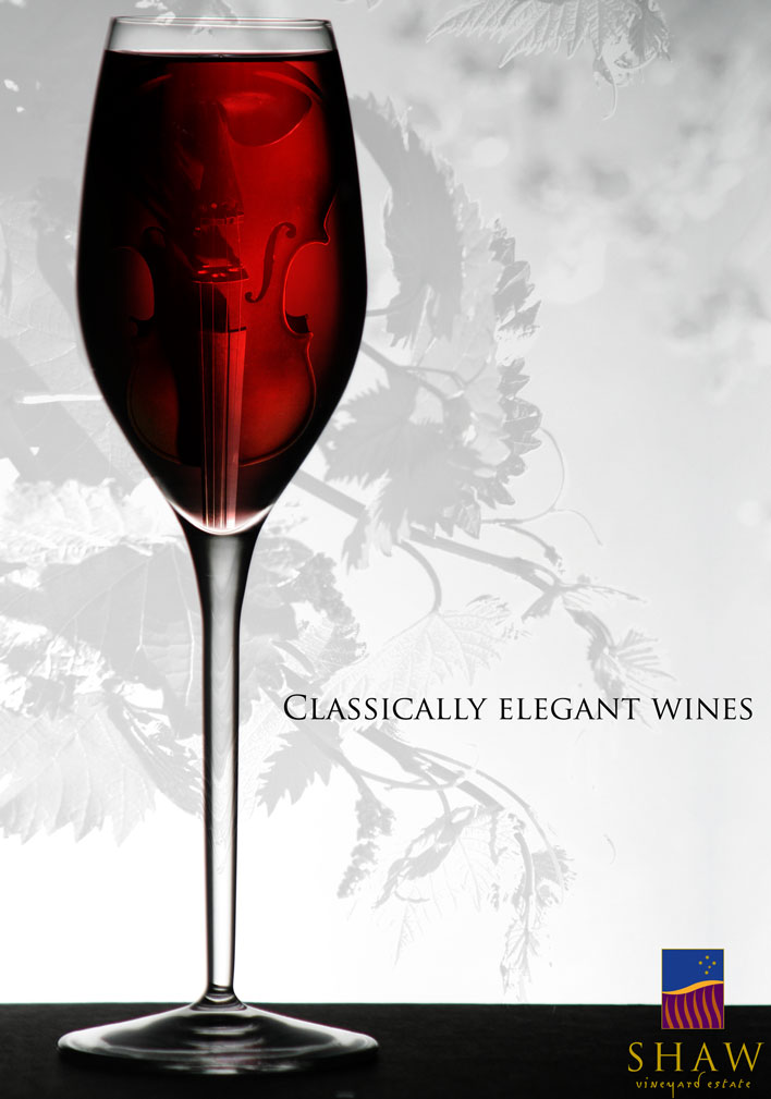 The poster for Shaw Vineyard Estate features an elegant Italian-made wine glass filled with red wine. Within the shadows of the wine glass a violin can be seen. The striking colour of the wine stands out against the soft black and white photograph of grapevines in the background.